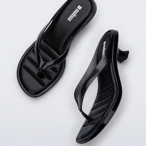 May include: A pair of black thong sandals with a low kitten heel and a glossy finish. The footbed features a quilted texture and the brand name melissa is printed in white on the heel area. The sandals have a rounded toe and a slim strap design.