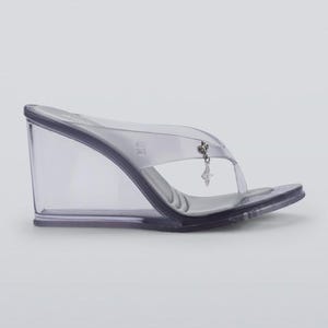 Könnte beinhalten: Transparente Keilsandalen mit Zehensteg. Der transparente Keilabsatz hat eine sanfte Kurve und einen kleinen Anhänger. Die Sandalen bestehen aus transparentem Material und haben ein minimalistisches Design. Absatzhöhe ca. 10 cm.