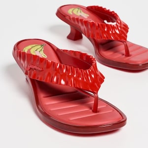 Pode incluir: Sandálias vermelhas estilo chinelo com uma tira com babados e salto pequeno. A palmilha tem um design texturizado. Um gráfico de banana amarela e verde é visível na palmilha. As sandálias são brilhantes.
