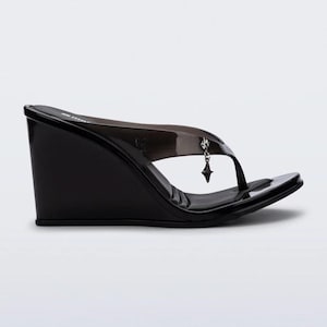Könnte beinhalten: Schwarze Keilsandalen mit transparentem Riemen und kleinem Anhänger. Die Sandalen haben eine eckige Zehenpartie und einen Keilabsatz. Das Design ist schlicht und modern, passend für verschiedene Anlässe.