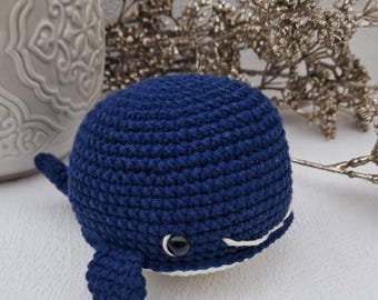 May include: A handmade, navy blue crochet whale with a white belly and a smiling mouth. The whale has small fins and a tail. The background includes a decorative vase and dried floral accents.