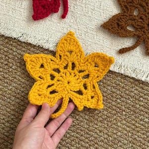 Puede incluir: Decoraciones de hojas de ganchillo en rojo, marrón y amarillo. Las hojas están dispuestas sobre una superficie texturizada. Una mano sostiene la hoja amarilla. Las hojas tienen un diseño calado detallado.