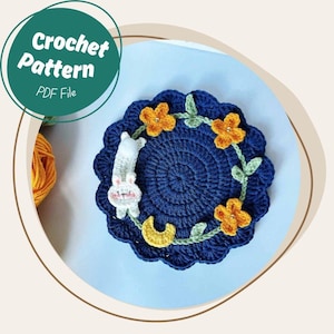 May include: A navy blue crochet coaster featuring a floral design and a white bunny. The coaster has a scalloped edge and a yellow crescent moon. The text "Crochet Pattern PDF File" is in a green circle.