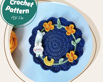 May include: A navy blue crochet coaster with a floral design and a white bunny. The coaster has a scalloped edge and a yellow crescent moon. The text "Crochet Pattern PDF File" is in a green circle.