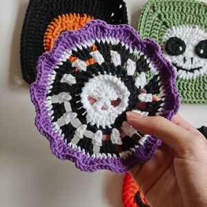 May include: A crocheted Halloween coaster with a skull design in white, black, and purple. The coaster is held in front of other crocheted Halloween-themed squares, including a black square with orange accents and a green square with a smiling face.