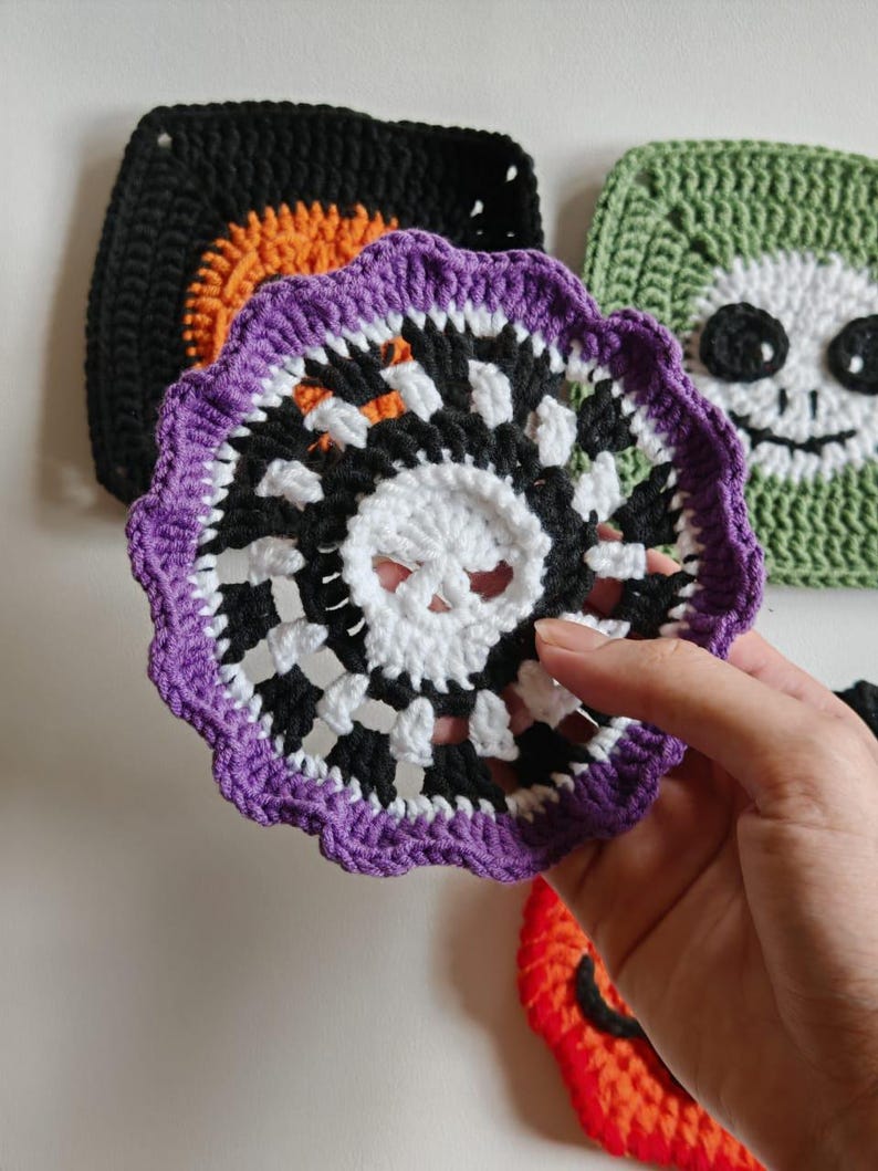 May include: A crocheted Halloween coaster with a skull design in white, black, and purple. The coaster is held in front of other crocheted Halloween-themed squares, including a black square with orange accents and a green square with a smiling face.
