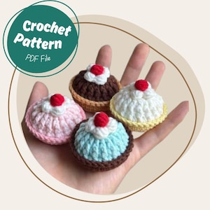 Könnte beinhalten: Vier Miniatur-Häkel-Cupcakes in Rosa, Braun, Blau und Weiß, jeder mit einer roten Kirsche belegt. Die Cupcakes werden in einer Hand gehalten. Das Bild enthält einen grünen Kreis mit dem Text "Crochet Pattern PDF File."