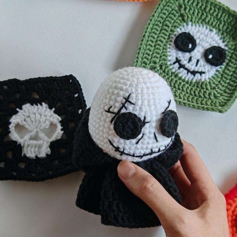 May include: A white crocheted skull doll with black eyes and stitched details, wearing a black cloak. Beside it are crocheted squares with skull faces in black and white, and green and white. The doll is held by a hand.