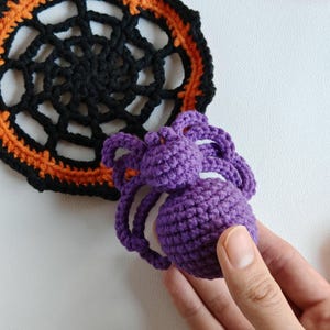 May include: A crocheted Halloween spider in purple, held by a hand, next to a black and orange crocheted spiderweb doily. The spider has a round body and eight legs. The doily is circular with a web design.