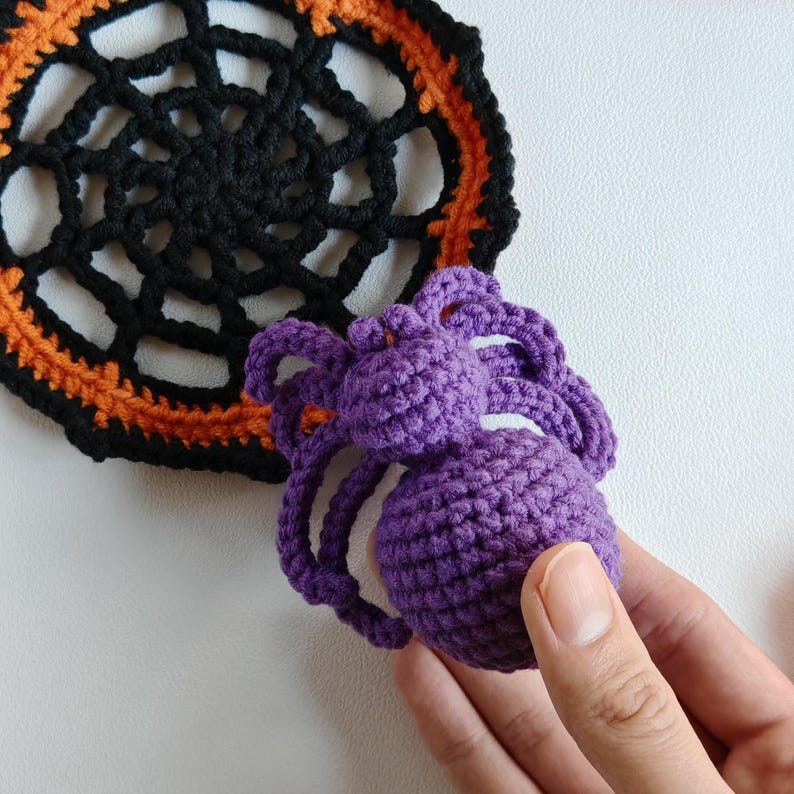 May include: A crocheted Halloween spider in purple, held by a hand, next to a black and orange crocheted spiderweb doily. The spider has a round body and eight legs. The doily is circular with a web design.