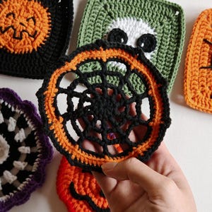 May include: Handmade crochet Halloween coasters in various designs. The coasters feature a black spiderweb design with orange trim, a green square with a white skull, and orange jack-o'-lantern faces. The coasters are made of yarn.