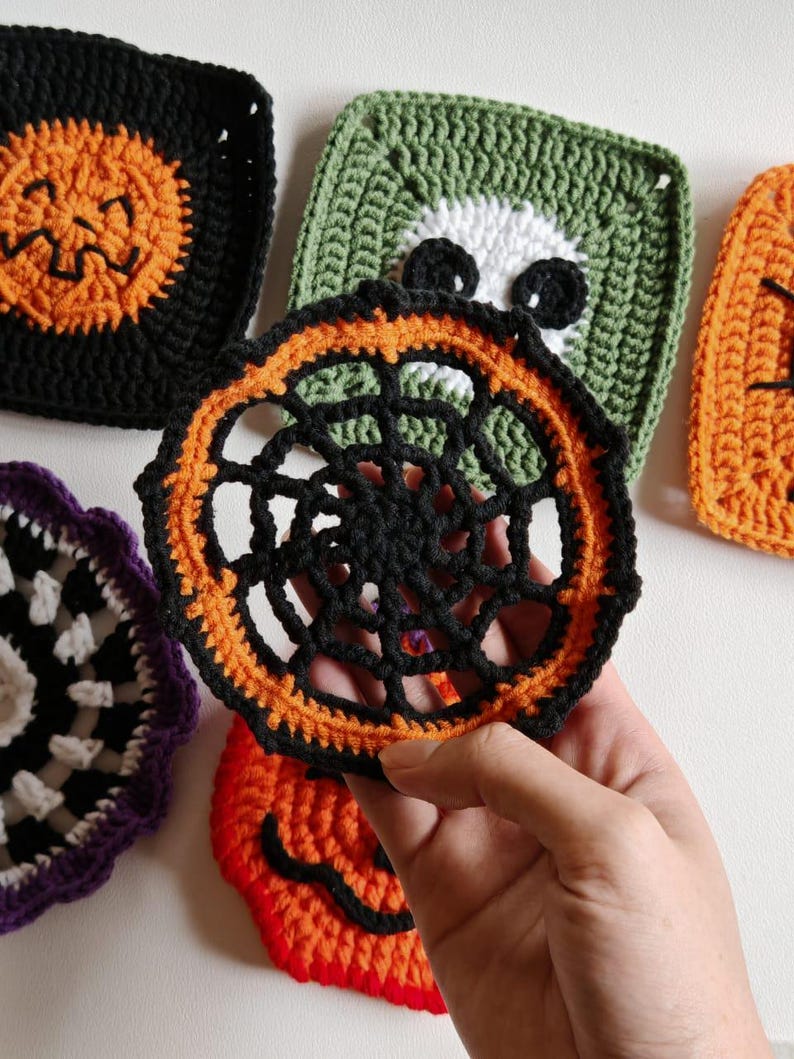May include: Handmade crochet Halloween coasters in various designs. The coasters feature a black spiderweb design with orange trim, a green square with a white skull, and orange jack-o'-lantern faces. The coasters are made of yarn.