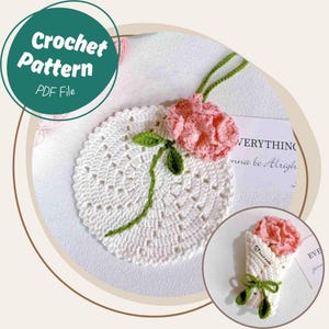 Puede incluir: Un posavasos blanco de ganchillo con una flor rosa de ganchillo y un tallo verde. La imagen también incluye un ramo de flores de ganchillo más pequeño. El texto "Crochet Pattern PDF File" está en un círculo verde.