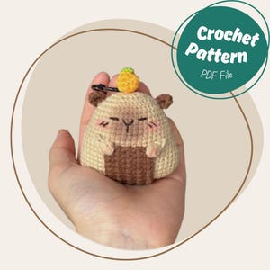 May include: A crocheted hamster toy with a small pineapple on its head. The hamster is beige and brown with a smiling face and small arms. The image includes a green circle with the text "Crochet Pattern PDF File".
