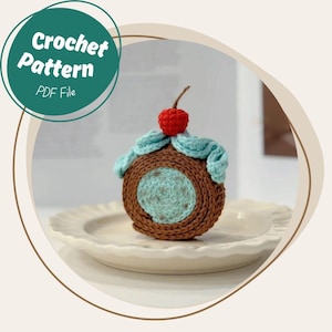 May include: A crocheted dessert, resembling a cupcake or pastry, with a brown exterior, a light blue center, and a red cherry on top. The dessert sits on a white plate. The image includes a circular graphic with the text "Crochet Pattern PDF File."