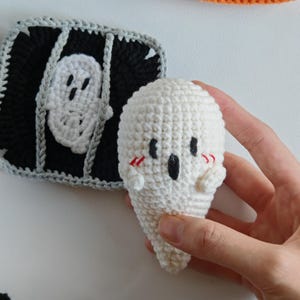 May include: A white crochet ghost with black eyes, a black mouth, and red blush marks is held in a hand. Beside it is a black and gray crochet square with a white ghost design.