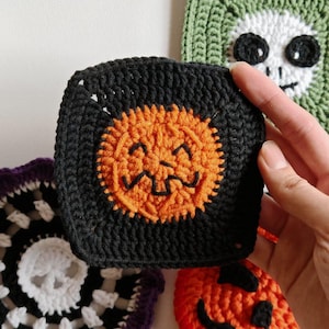 May include: A collection of handmade crochet Halloween-themed squares. The squares feature a black square with an orange jack-o'-lantern face, a green square with a white skull, and a red and orange pumpkin face.