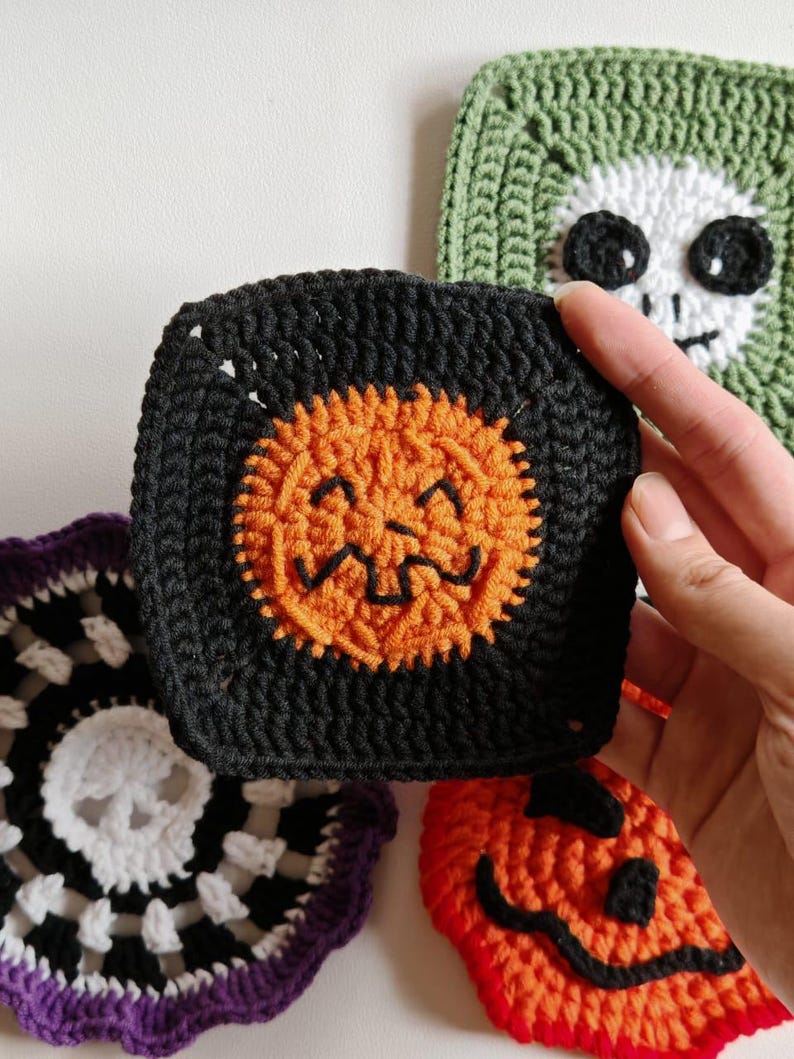 May include: A collection of handmade crochet Halloween-themed squares. The squares feature a black square with an orange jack-o'-lantern face, a green square with a white skull, and a red and orange pumpkin face.