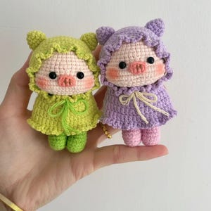 May include: Two handmade crochet pig dolls. One doll is lime green, the other is lavender. Both dolls have pink snouts, rosy cheeks, and black eyes. They wear hooded capes with ruffled edges and are tied with a bow.