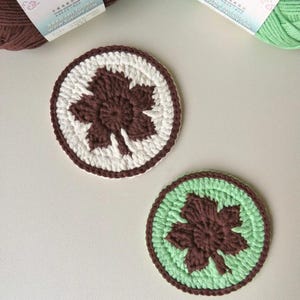 May include: Two handmade crochet coasters. One coaster features a brown maple leaf design on a white background, framed by a brown border. The other coaster has a brown maple leaf on a light green background, with a brown border. Yarn skeins are in the background.
