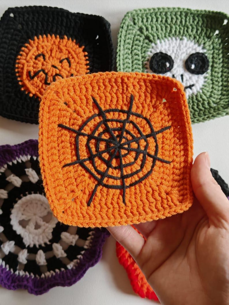 May include: Close-up of several crocheted Halloween-themed squares. One orange square features a black spiderweb design. Other squares include a black pumpkin, a green skull, and a purple and white design. The squares are handmade.