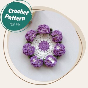 May include: A handmade crochet pattern featuring a floral design. The pattern includes a central white and purple flower with surrounding purple flowers. The text "Crochet Pattern PDF File" is in a green circle.