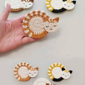 May include: Five crocheted cat coasters in different color combinations. The coasters are shaped like sleeping cats with white, brown, and yellow yarn.
