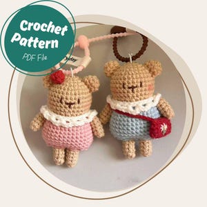 May include: Two crocheted bear keychains. One bear wears a pink dress and a red flower, the other wears a blue dress and a red purse. The keychains are tan, with white collars. The image includes the text "Crochet Pattern PDF File".