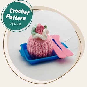 May include: A pink crocheted cupcake with a white and pink floral design, sitting on a blue tray with two pink plastic utensils. The image includes a green circle with the text "Crochet Pattern PDF File".