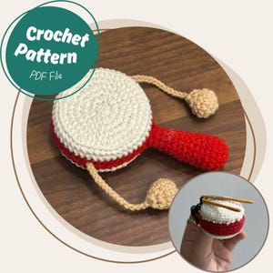 Puede incluir: Un juguete de tambor de ganchillo hecho a mano con la parte superior blanca, los lados y el mango rojos y los detalles en beige. El texto "Crochet Pattern PDF File" está en un círculo verde. El tambor tiene brazos beige y manos en forma de bola beige.
