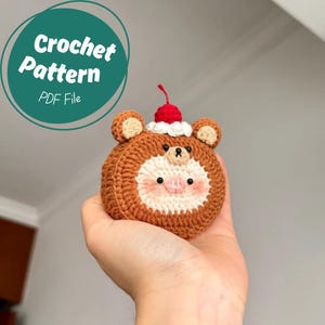 May include: A crocheted bear-shaped ornament with a cherry on top. The bear has brown fur, a white face, and small black eyes. The text "Crochet Pattern PDF File" is in a green circle.