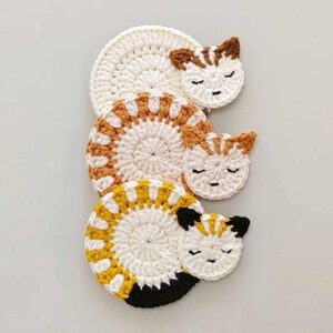May include: Set of three crocheted cat-shaped coasters in white, yellow, and orange with black accents. The coasters are stacked on top of each other.