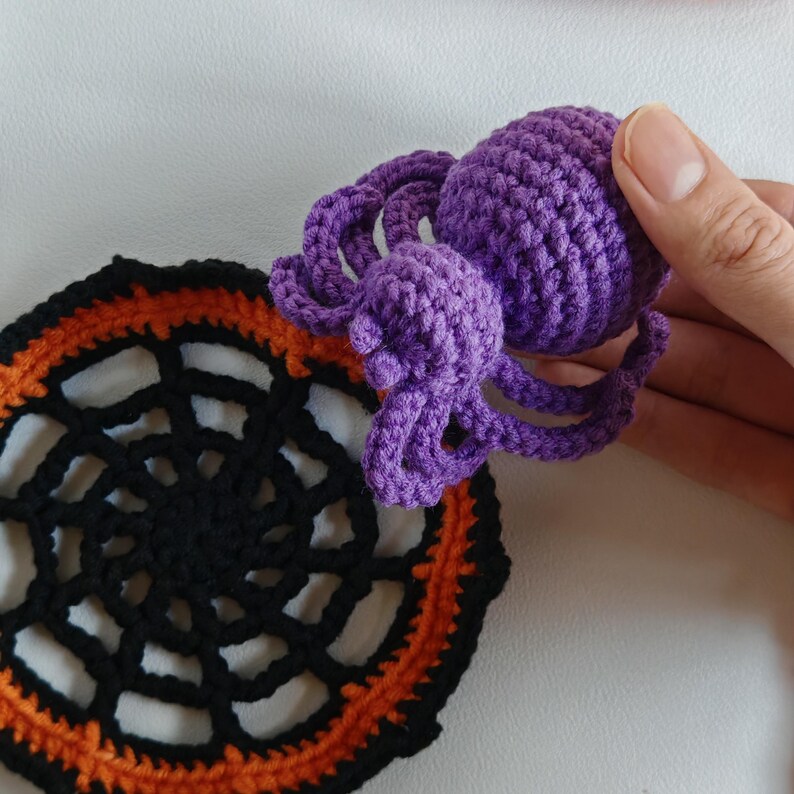 May include: A crocheted Halloween-themed set featuring a purple spider and a black and orange spiderweb coaster. The spider has a rounded body and eight curled legs. The coaster is round with a web design and orange trim.