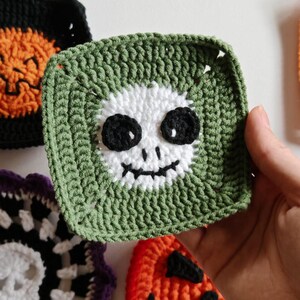 May include: Crocheted Halloween-themed squares in various colors. A green square features a white skull face with black eyes and a mouth. Other squares include a black and orange pumpkin, and a black and white skull design. These are likely coasters or decorative items.
