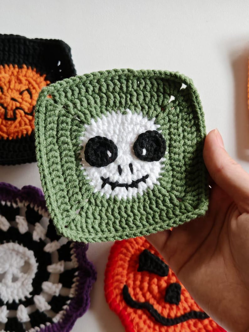 May include: Crocheted Halloween-themed squares in various colors. A green square features a white skull face with black eyes and a mouth. Other squares include a black and orange pumpkin, and a black and white skull design. These are likely coasters or decorative items.