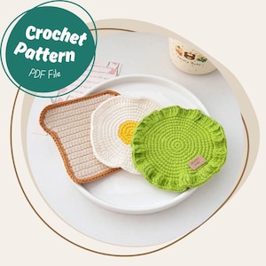 Puede incluir: Un conjunto de tres posavasos de ganchillo con temática de comida: una rebanada de pan tostado, un huevo frito y un posavasos verde con un borde con volantes. El posavasos de pan tostado es de color beige con un borde marrón. El posavasos de huevo es blanco con un centro amarillo. El posavasos verde tiene una pequeña etiqueta que dice "Happy Baby". El texto "Crochet Pattern PDF File" está en un círculo verde.