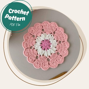 Pode incluir: Um descanso de copo de crochê rosa com um design de flor branca e rosa no centro. Arquivo PDF de padrão de crochê.
