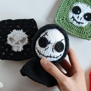 May include: Handmade crochet Halloween decorations. The image features a white and black crochet Grim Reaper doll with a black hood, and two crochet squares with skull faces. One square is black with a white skull, and the other is green with a white skull.