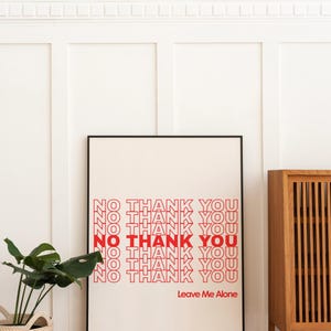 May include: A framed art print with the words "NO THANK YOU" repeated in red, with the phrase "Leave Me Alone" below. The print is displayed against a white wall, with a plant in a woven basket to the left.
