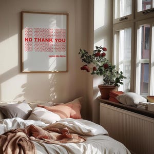 May include: A bedroom scene featuring a bed with white bedding, peach and gray pillows, and a brown blanket. A framed print on the wall reads "NO THANK YOU" in red text. A plant in a terracotta pot sits on the windowsill.