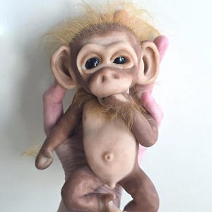 May include: A realistic monkey figurine with smooth, light brown skin, large dark eyes, and prominent ears. The figure features a tuft of wispy, golden-blonde hair on its head and chin, a small navel, and a hand positioned near its mouth.