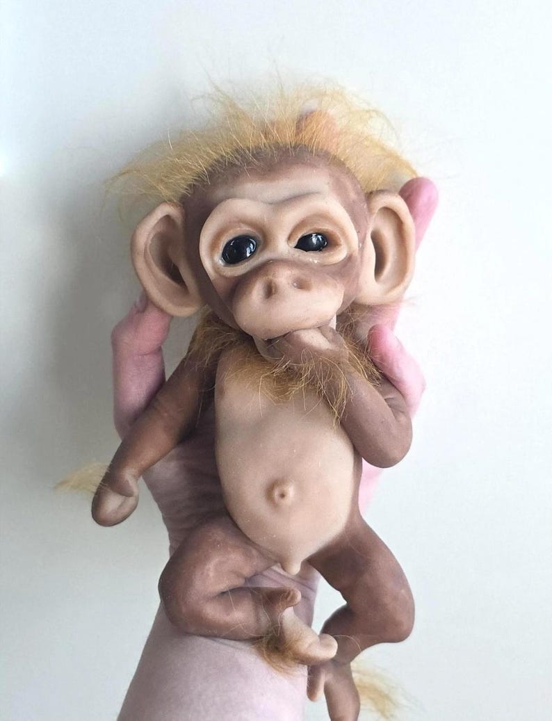 May include: A realistic monkey figurine with smooth, light brown skin, large dark eyes, and prominent ears. The figure features a tuft of wispy, golden-blonde hair on its head and chin, a small navel, and a hand positioned near its mouth.
