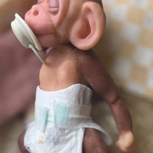 May include: A small, realistic monkey figurine with brown skin, large ears, and closed eyes. The monkey has a white pacifier in its mouth and is wearing a white disposable diaper featuring a small gray elephant graphic on the front panel.