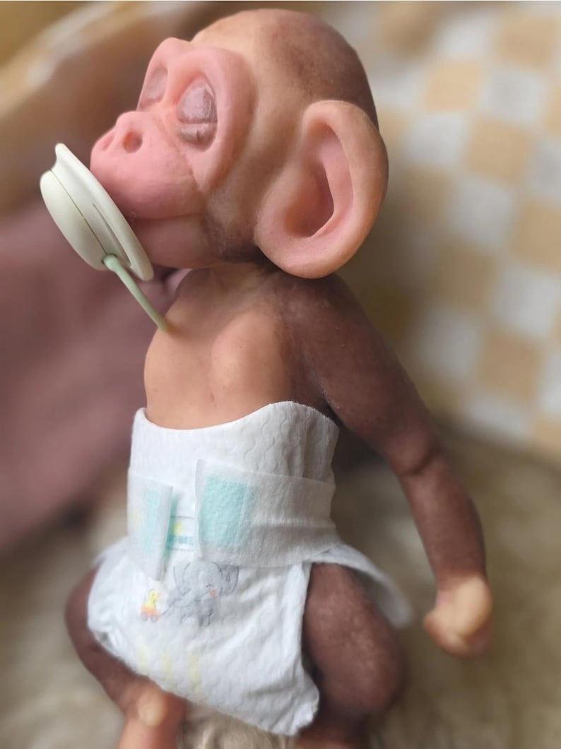 May include: A small, realistic monkey figurine with brown skin, large ears, and closed eyes. The monkey has a white pacifier in its mouth and is wearing a white disposable diaper featuring a small gray elephant graphic on the front panel.