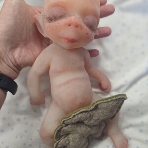 May include: A small, pale, fantasy-style doll with a smooth, skin-like texture, pointed ears, and long dark eyelashes. The doll has light freckles across the nose and cheeks, and is partially covered by a textured, leaf-shaped piece of grey-green felt.