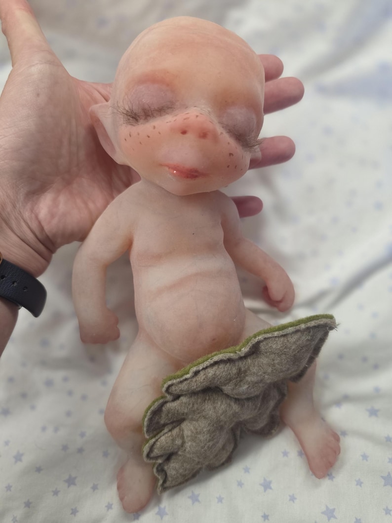 May include: A small, pale, fantasy-style doll with a smooth, skin-like texture, pointed ears, and long dark eyelashes. The doll has light freckles across the nose and cheeks, and is partially covered by a textured, leaf-shaped piece of grey-green felt.