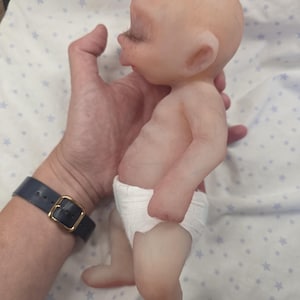 May include: A small, bald, light-skinned silicone doll with a rounded head, closed eyes, and visible eyelashes. The doll has a smooth, realistic skin texture, a slightly protruding belly, and is wearing a simple white diaper around its waist.
