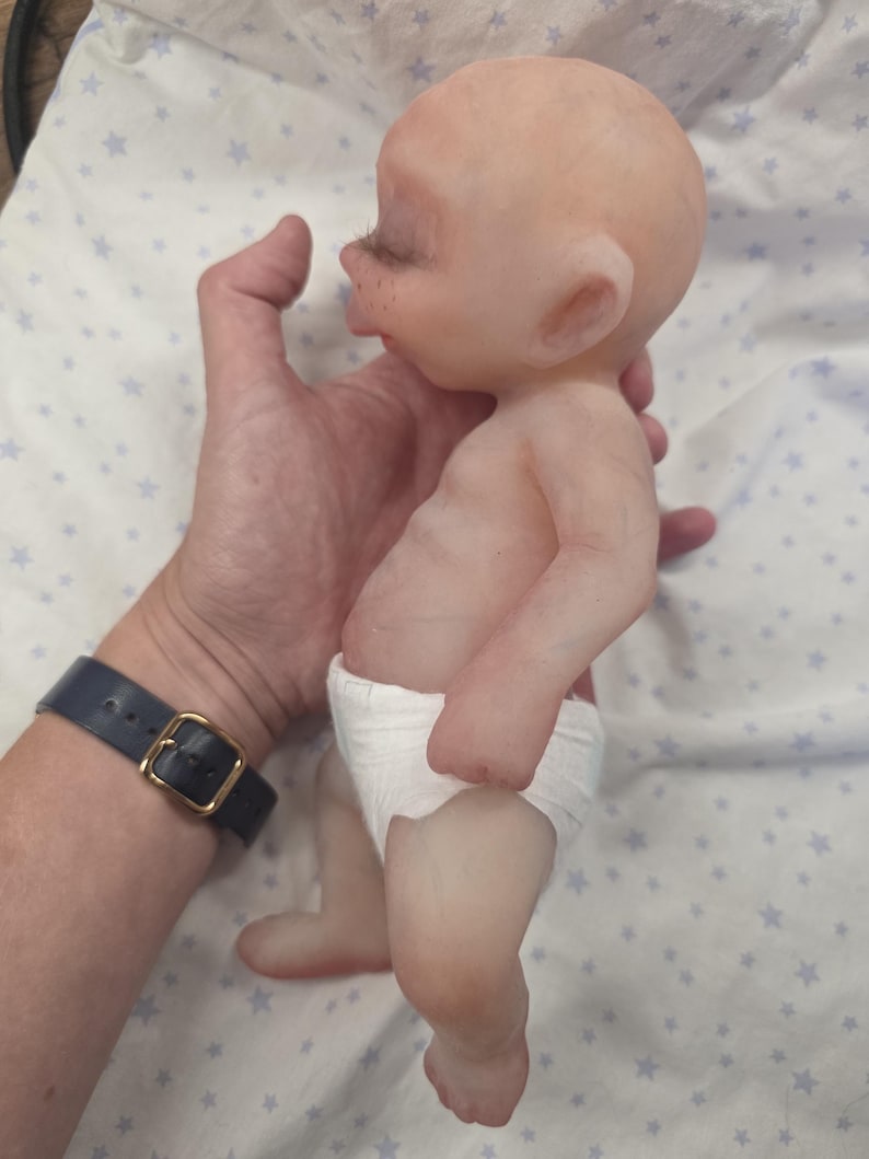 May include: A small, bald, light-skinned silicone doll with a rounded head, closed eyes, and visible eyelashes. The doll has a smooth, realistic skin texture, a slightly protruding belly, and is wearing a simple white diaper around its waist.