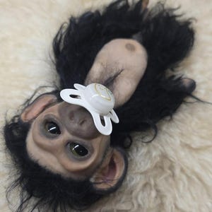 May include: A realistic chimpanzee doll with dark, textured fur, light-toned skin on the face and chest, and large, dark eyes. The doll features a white pacifier with a small yellow graphic design in its mouth, positioned against a soft, cream-colored surface.