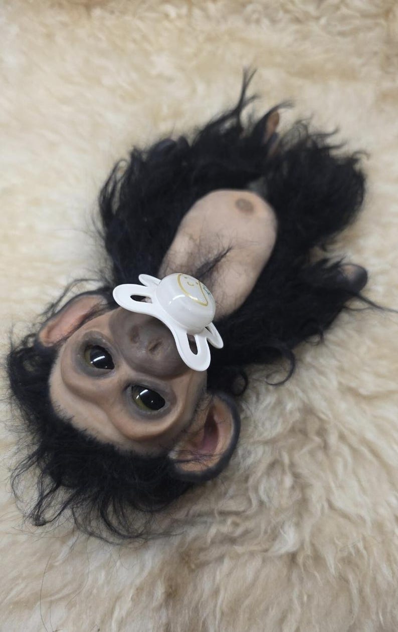 May include: A realistic chimpanzee doll with dark, textured fur, light-toned skin on the face and chest, and large, dark eyes. The doll features a white pacifier with a small yellow graphic design in its mouth, positioned against a soft, cream-colored surface.
